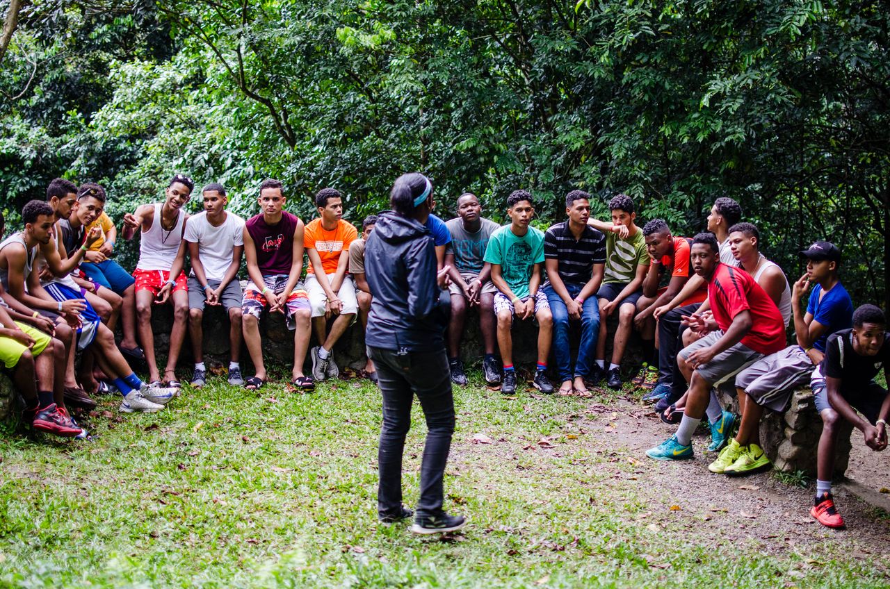 About A group of teenagers and adults listen attentively during an outdoor session in a green park.